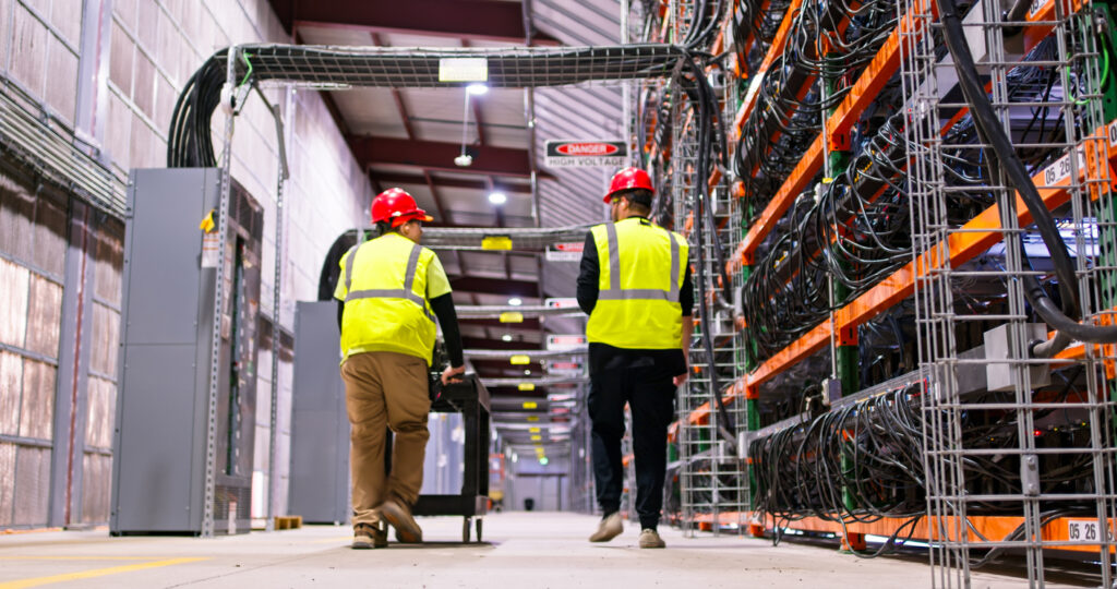 Two technicians walking past racks of equipment in a data center for cryptocurrency mining, cloud services and AI computing in a large, temperature controlled warehouse in a remote location in  Stutsman County, North Dakota.